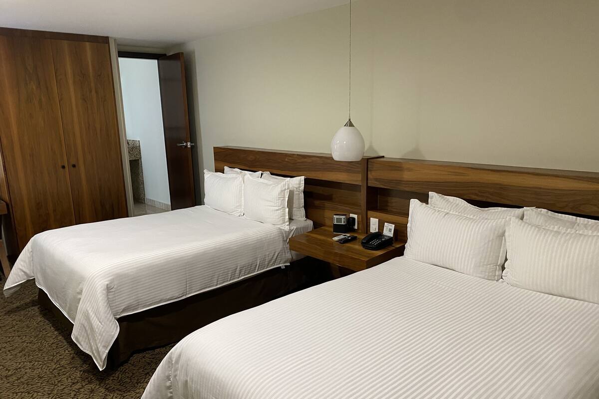 A hotel room with two single beds, a wooden headboard, a bedside table with a lamp, and a door leading to another room.