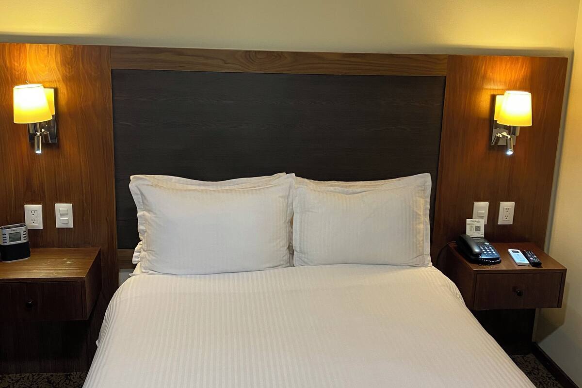 A hotel room with a large bed, two bedside tables with lamps, a wooden headboard, and a nightstand with a phone and remote control.