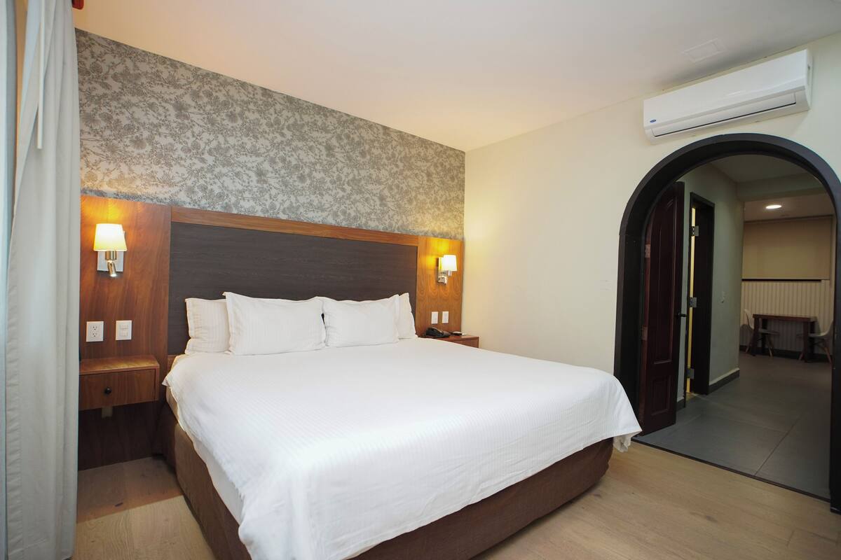 A hotel room with a large bed, wooden headboard, two bedside lamps, and an air conditioning unit.