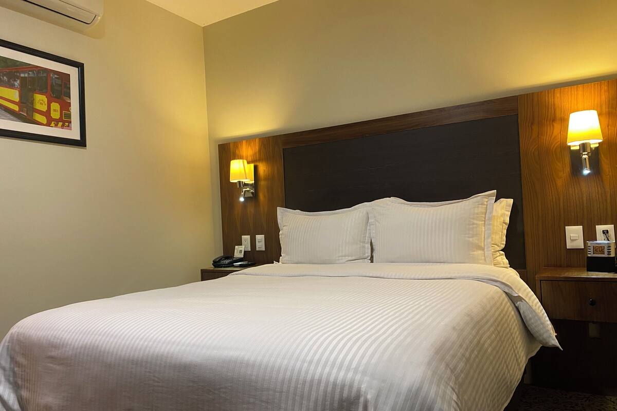 A hotel room with a large bed, two bedside tables, a wall-mounted lamp, a framed picture, and a clock.