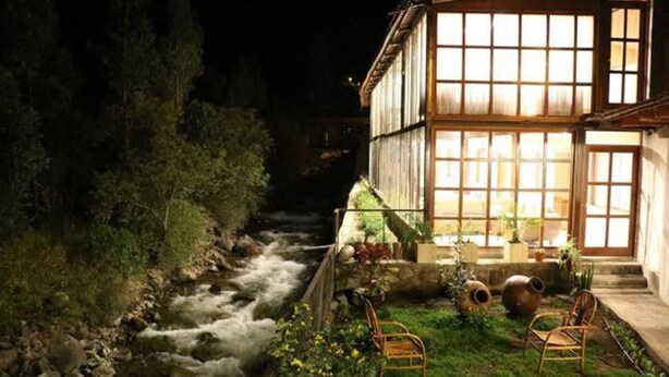 A riverside building with a well-lit entrance and a grassy area with chairs. - Hotel Humantay Lodge Ollantaytambo (Ollantaytambo)