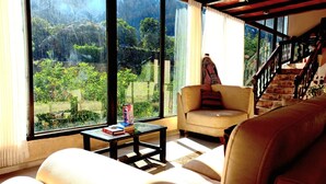 A living room with a sofa, a small table with books, and a chair. There is a staircase leading to an upper level. Large windows provide a view of greenery outside. - Hotel Humantay Lodge Ollantaytambo (Ollantaytambo)