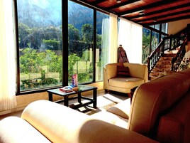 Salón con sofá, una mesita con libros y una silla. Una escalera conduce a un nivel superior. Amplias ventanas ofrecen vistas al exterior con vegetación.