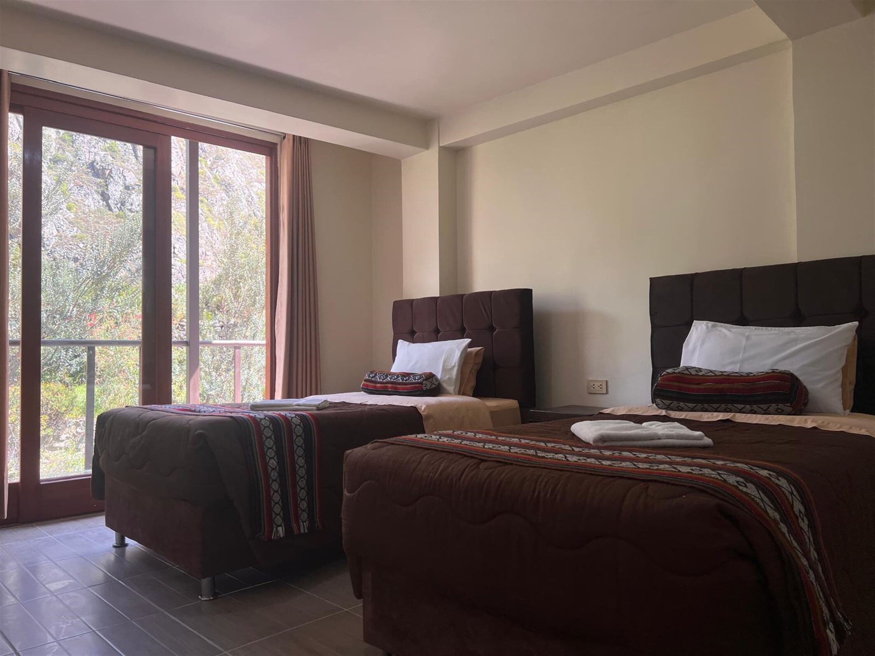 Habitación de hotel con dos camas, un ventanal con cortinas y vistas a árboles.
