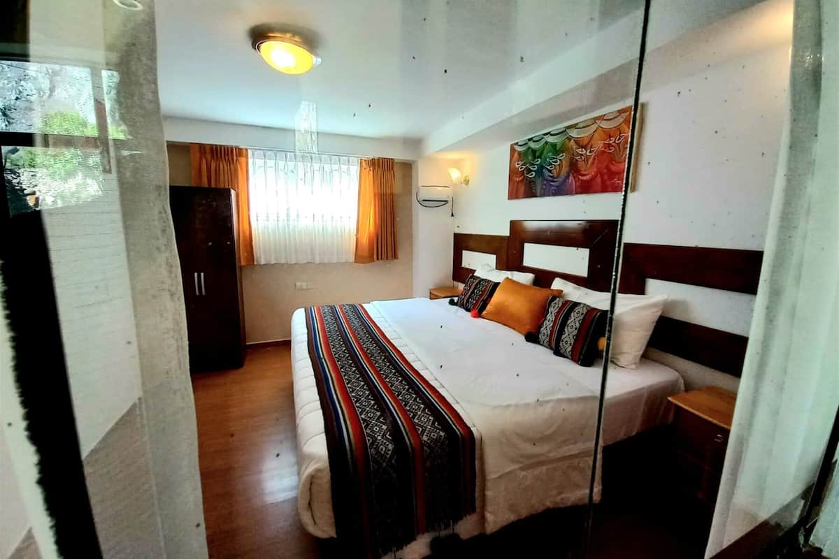 A hotel room with a bed, a wardrobe, a nightstand, a wall-mounted lamp, and a painting.