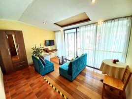 Un salón con suelo de madera, un sofá azul, un televisor montado en la pared y un amplio ventanal con cortinas.