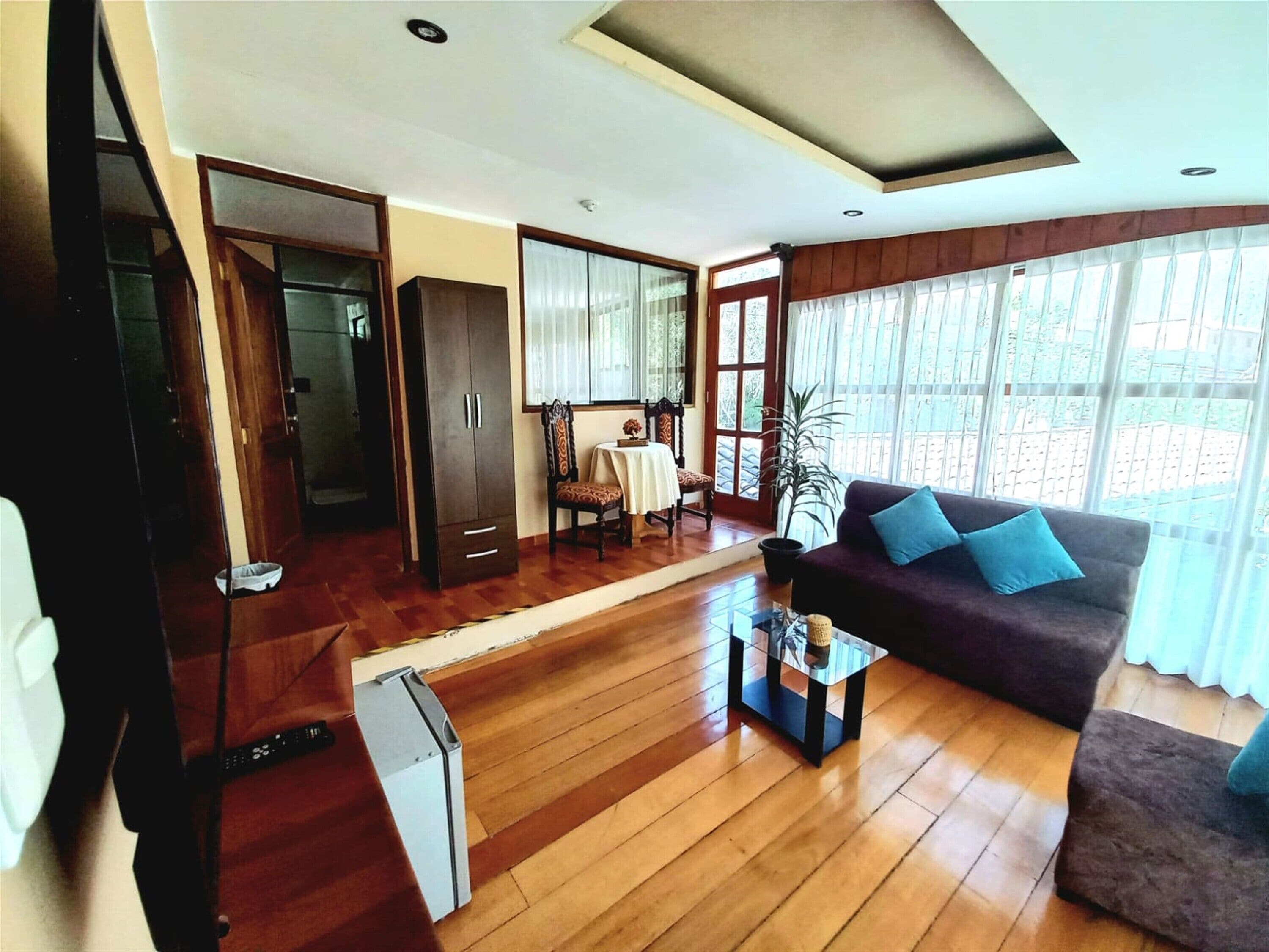 A spacious living area with wooden flooring, a sofa, a dining table, and a large window with curtains.