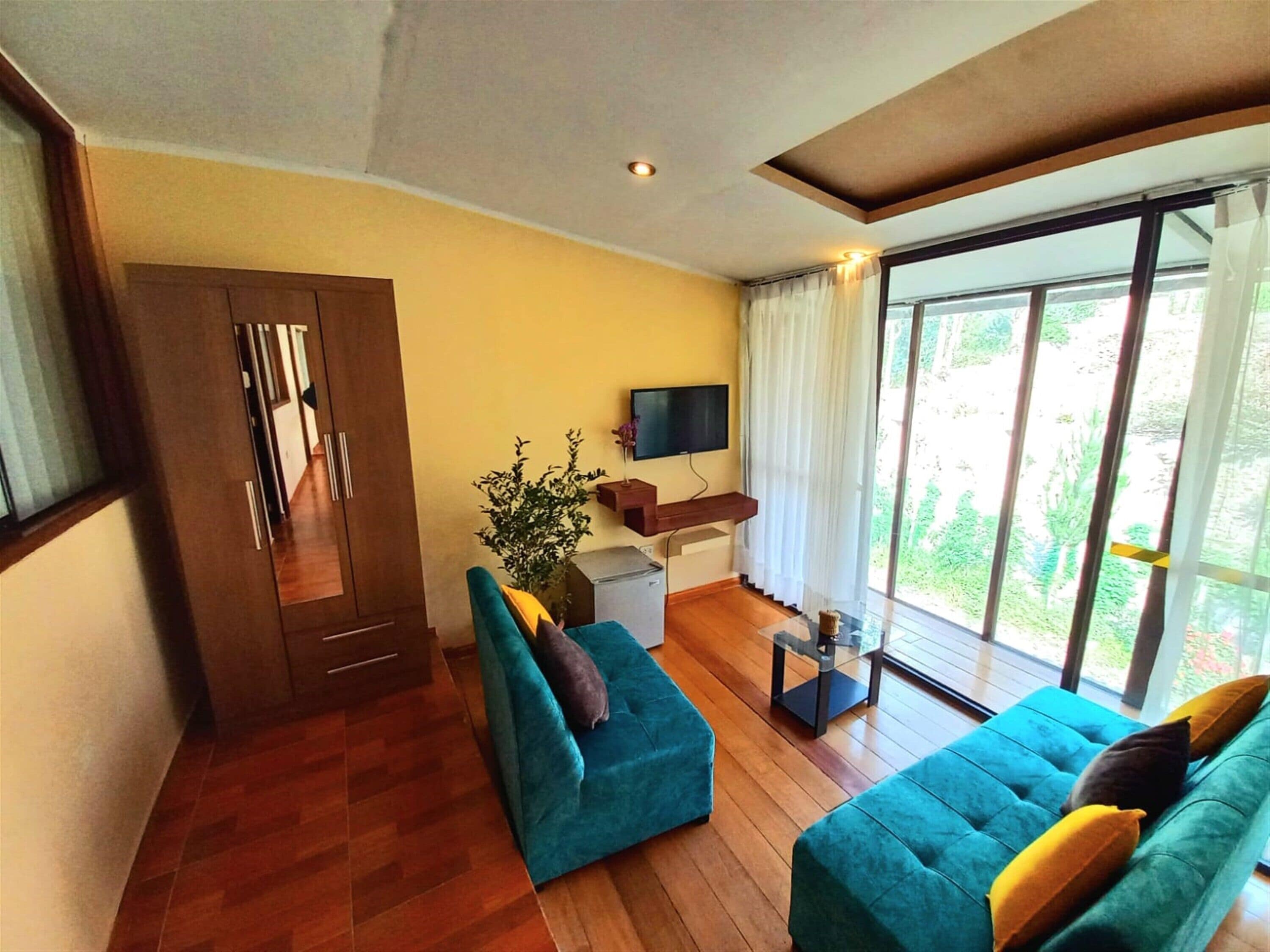 Uma sala de estar com piso de madeira, um sofá azul, uma televisão na parede e uma janela grande com cortinas.
