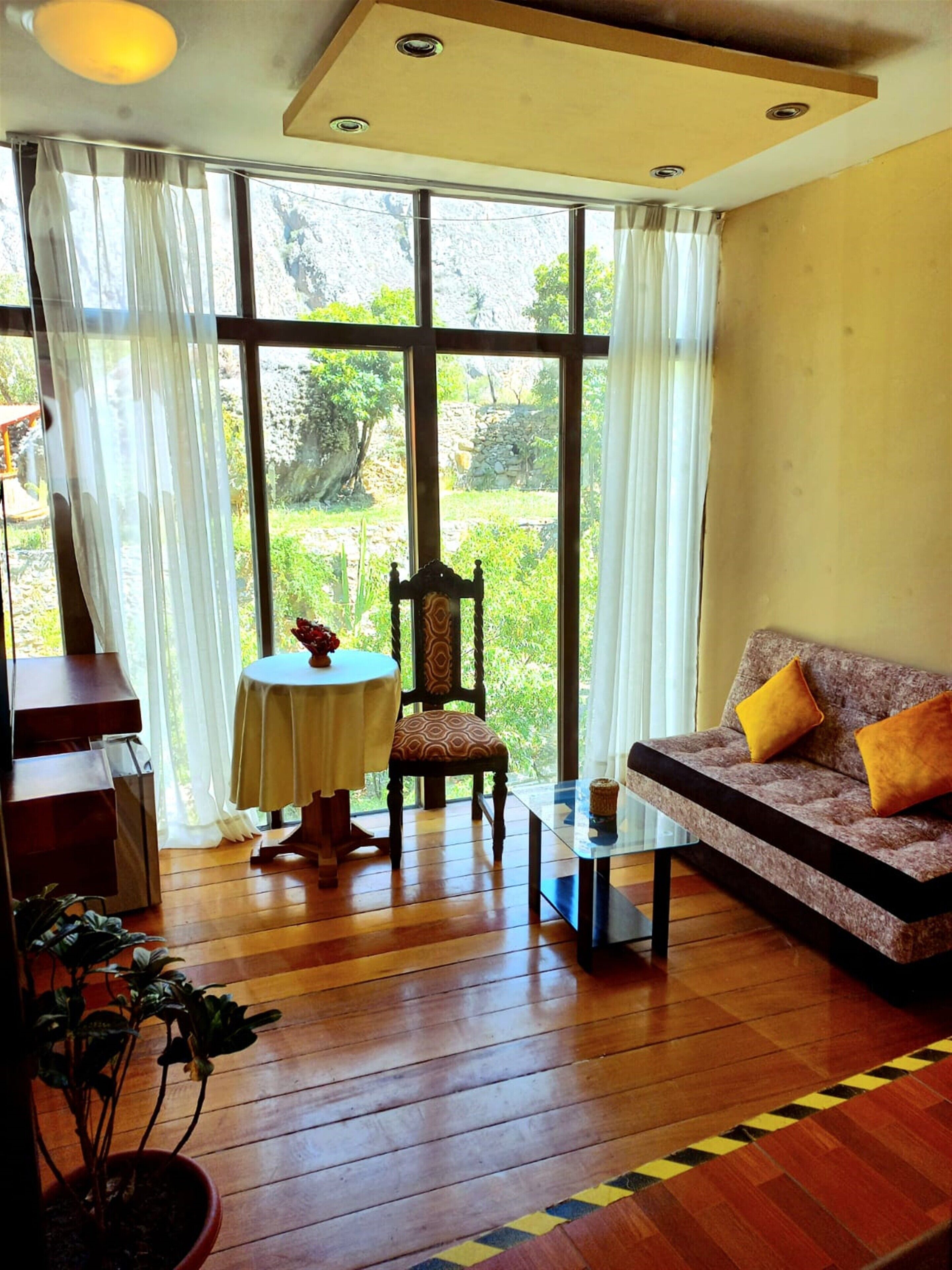 A room with a wooden floor, a sofa with yellow cushions, a chair, a small table, and a window with curtains.