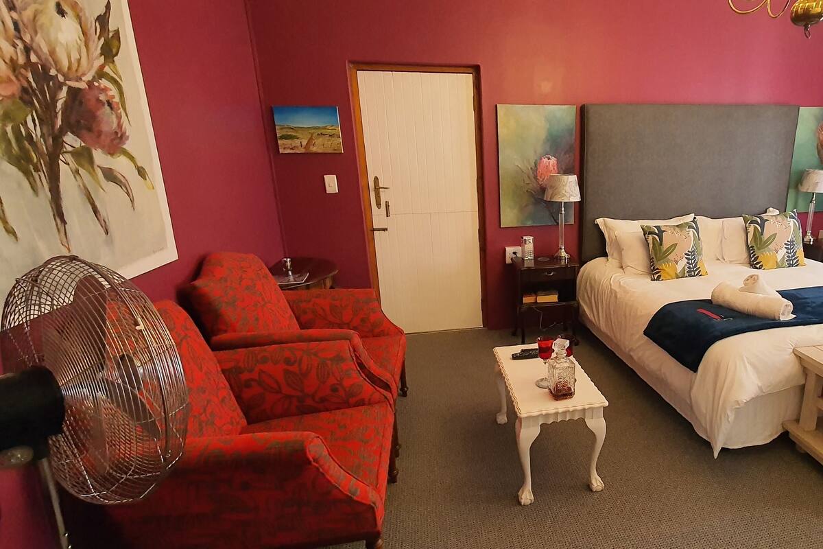 A hotel room with a bed, a red armchair, a small table, a fan, and artwork on the walls.