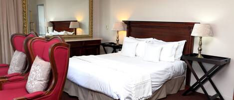 A hotel room with a large bed, two bedside tables, a mirror, a red armchair, and a wooden headboard.