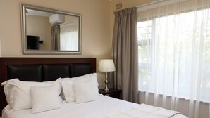 Een netjes opgemaakt bed met witte lakens en een decoratieve plaid, een bedlamp, een grote spiegel en lichte gordijnen voor het raam.