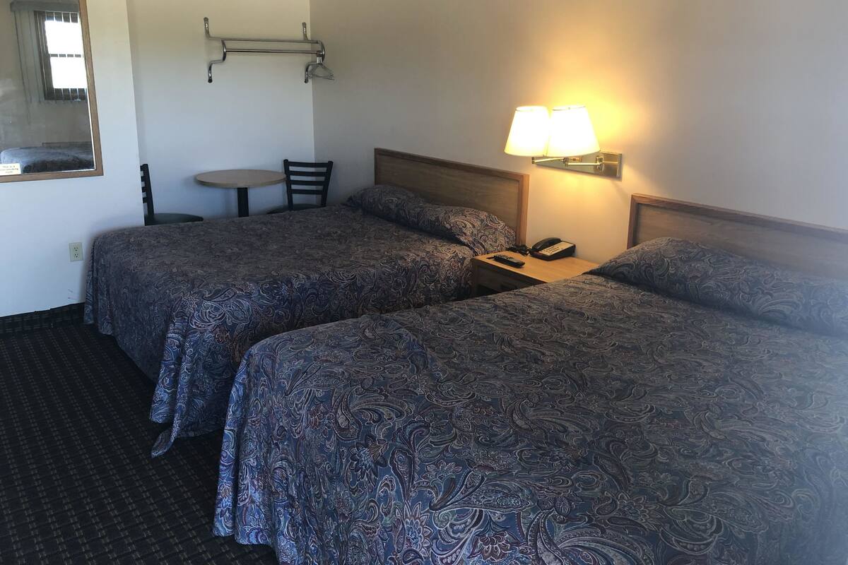A hotel room with two beds, a small table, a chair, a lamp, and a phone on the nightstand.
