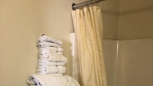 A bathroom with a stack of white towels on a wall-mounted rack, a beige shower curtain, and a shower stall. - Quik Stop and Motel (Wausaukee)