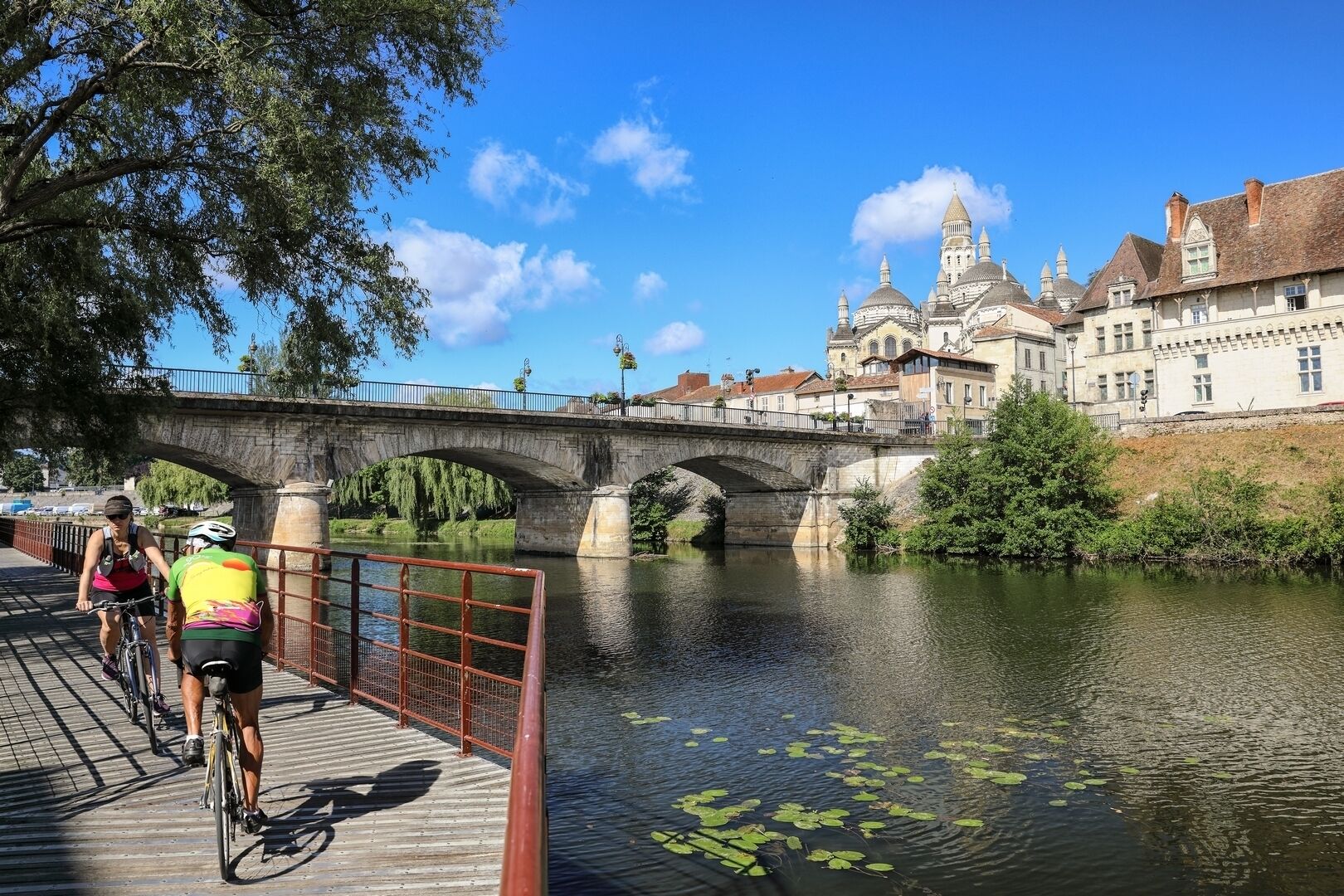 A bridge over a river with cyclists on a wooden walkway, a historic building with domes in the background.