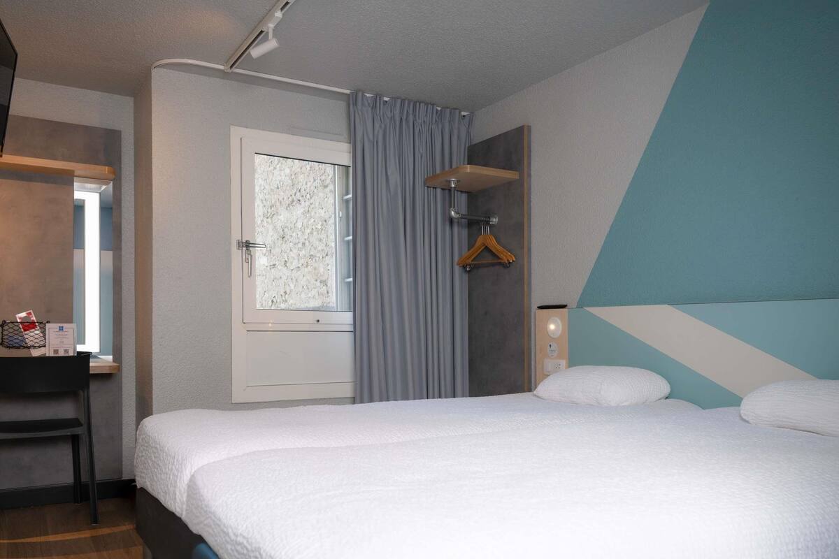A hotel room with a bed, a desk, a window with curtains, a wall-mounted light, and a geometric-patterned wall.
