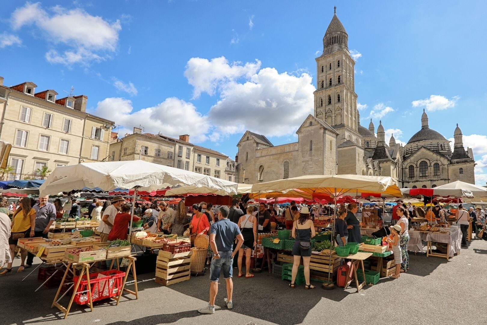 A bustling outdoor market with vendors and customers, set against historic buildings and a prominent tower.