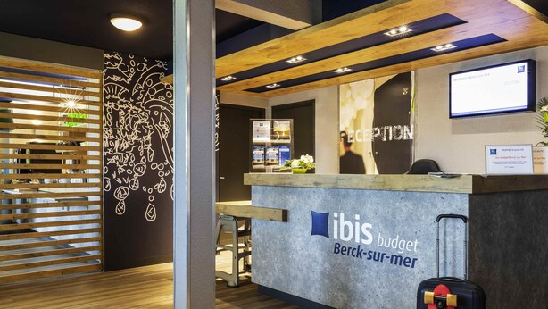 Un comptoir d'accueil avec l'enseigne « ibis budget », un paravent en bois à lattes et un téléviseur fixé au mur.