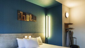 Une chambre d’hôtel avec un lit, des oreillers, un chapeau, des lunettes, un téléphone et un luminaire fixé au mur.