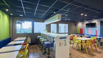Eine moderne Cafeteria mit hohen Tischen und bunten Stühlen, einer Theke mit einem QR-Code und großen Fenstern.