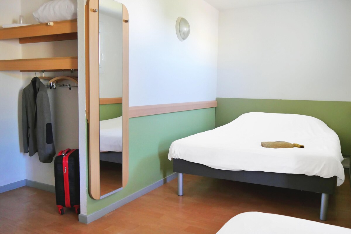 Une pièce avec un lit, une valise, un manteau et un miroir.