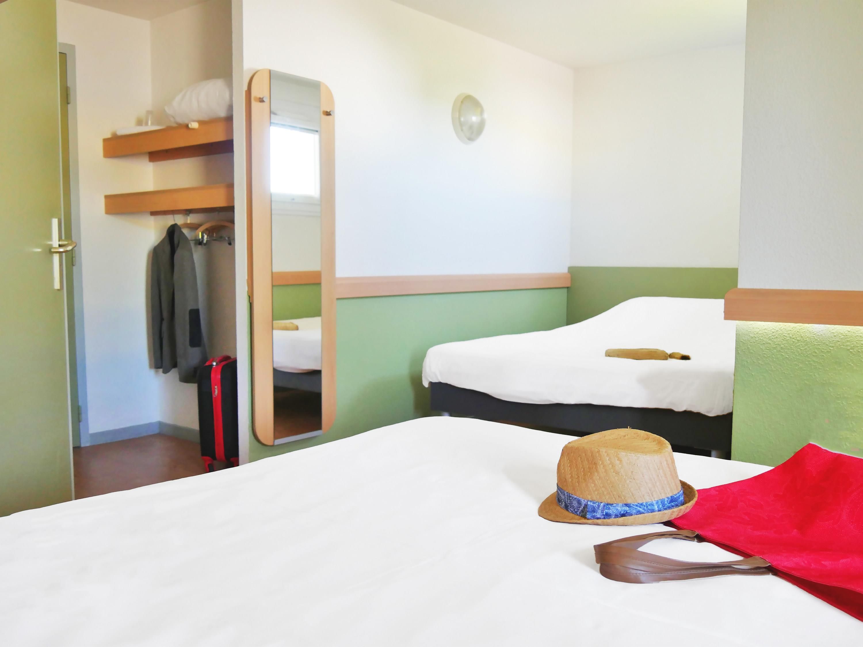 Ett hotellrum med en säng, en spegel, en resväska och en hatt som ligger på sängen.