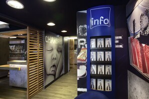 Een modern ingerichte receptiebalie met een receptiemedewerker, een muur met een groot poster en een blauwe informatiezuil met foto's.