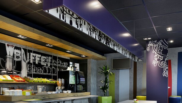 Een modern café met een lange toonbank, gele stoelen en een ruim assortiment aan eten en drinken.