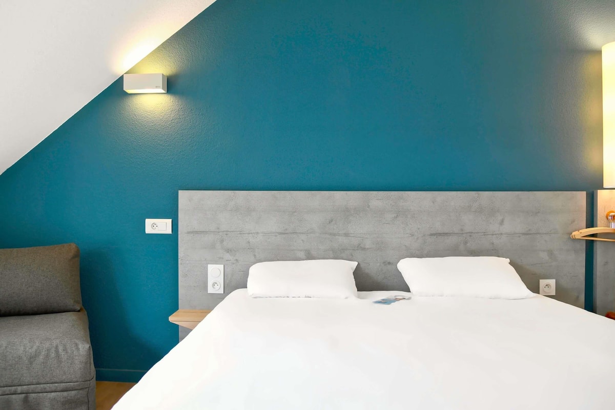Une chambre d’hôtel moderne avec un mur d’accent de couleur turquoise, une tête de lit grise et un lit blanc.