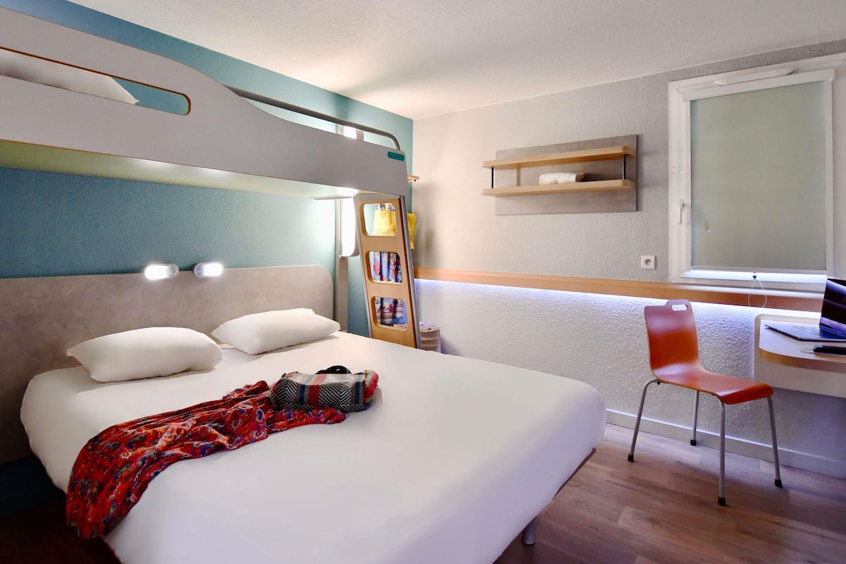 Une chambre d’hôtel avec un lit, un bureau avec un ordinateur portable, une chaise et une fenêtre.