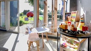 Une chambre moderne avec une porte vitrée donnant sur un jardin, une table blanche, une chaise en bois et une étagère remplie de jouets.