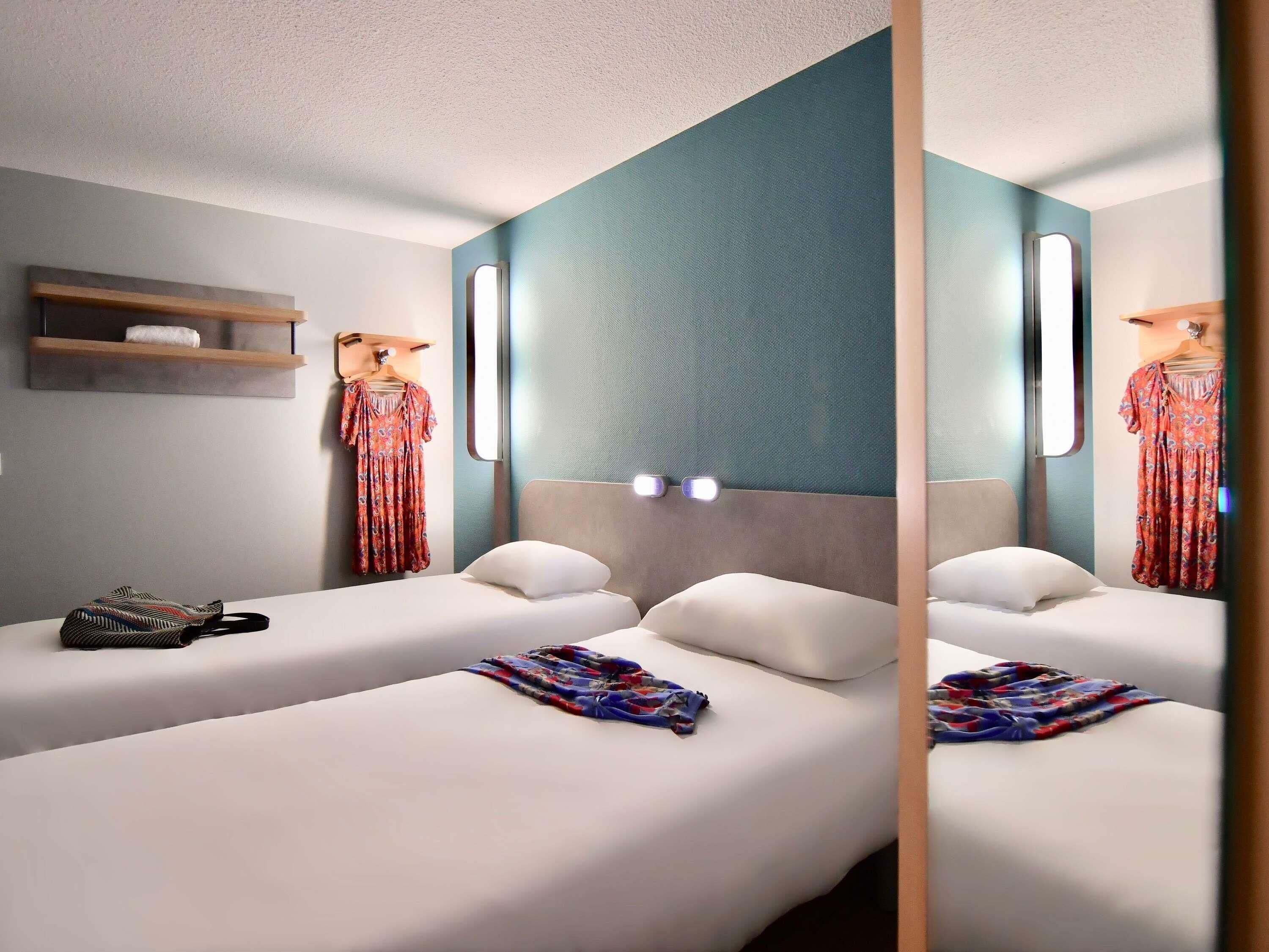 Habitación de hotel con dos camas, un estante de madera pequeño y un espejo que refleja un vestido.