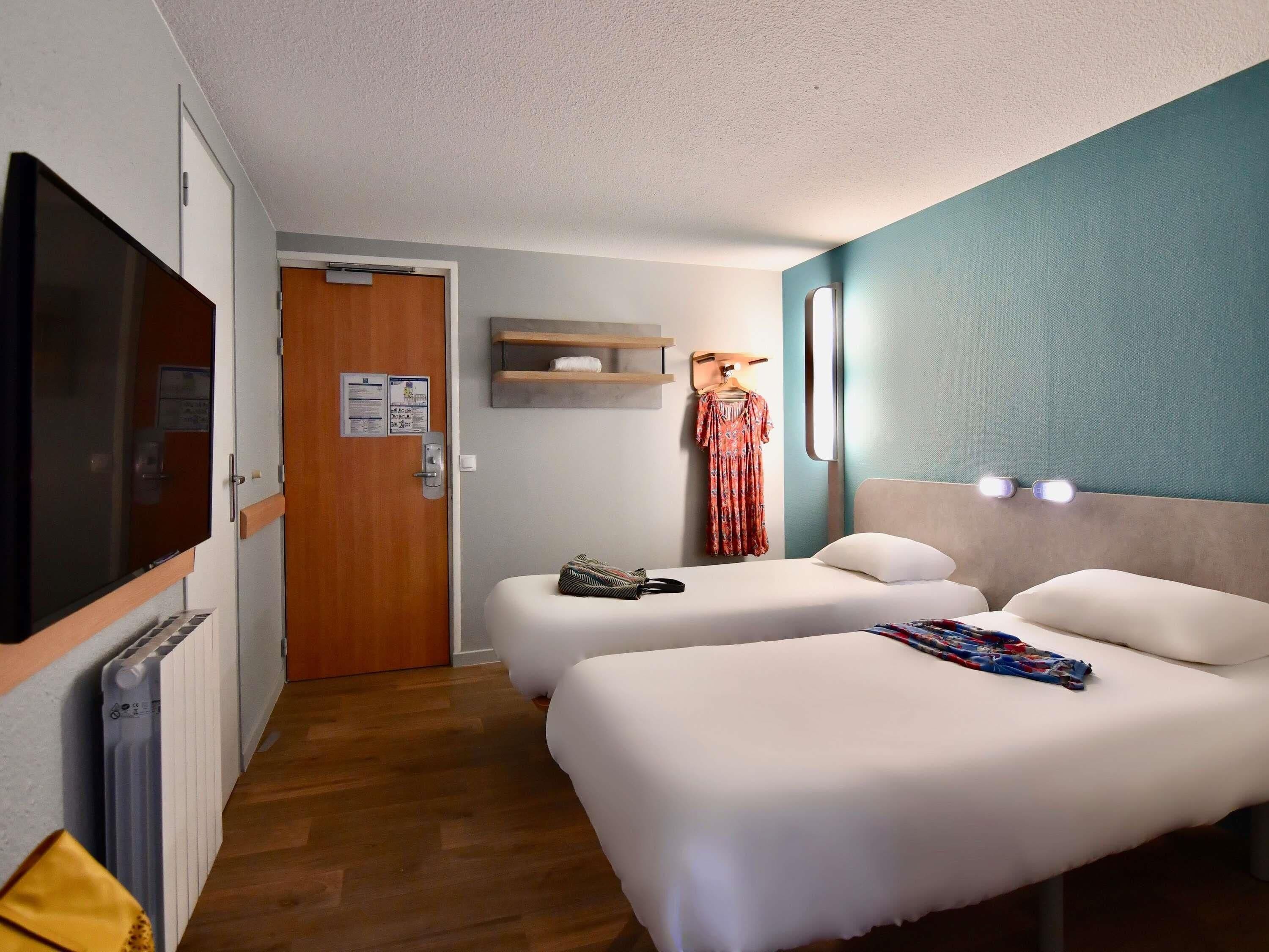 A hotel room with two beds, a TV, a wooden door, a towel rack, and a wall-mounted shelf.