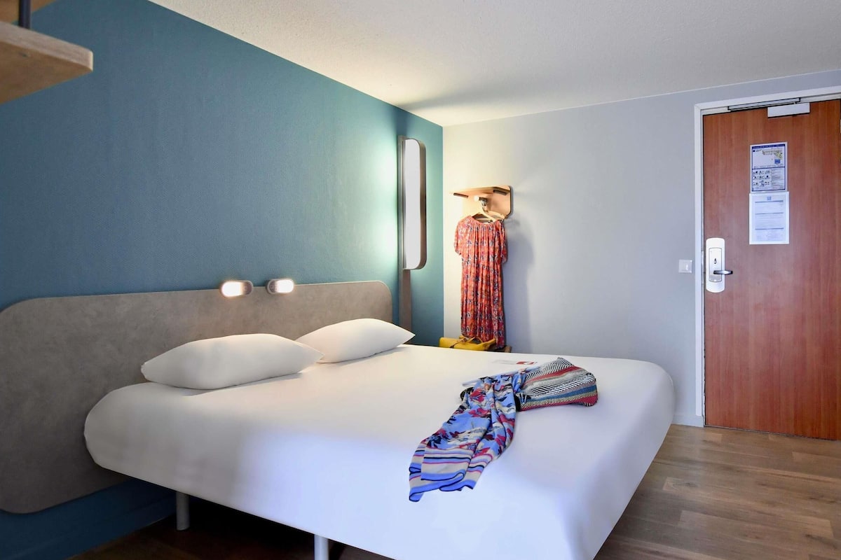 Un lit simple avec une literie blanche, une tête de lit dotée de lampes de lecture intégrées, une porte en bois avec un panneau d’affichage et un mur d’accent bleu.