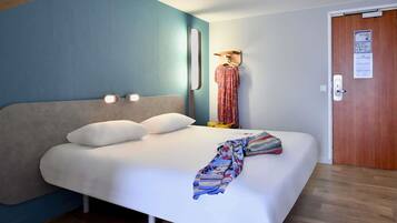Una cama individual con ropa de cama blanca, cabecera con luces de lectura incorporadas, una puerta de madera con un tablón de anuncios y una pared de acento azul.