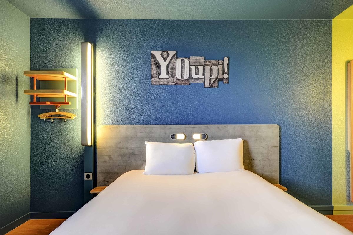 A bed with white linens, a headboard with two light switches, and a wall-mounted sign that reads 'Youpi!'.