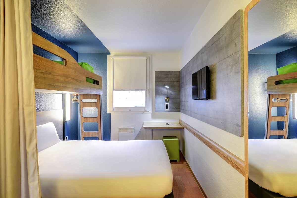 A compact hotel room with bunk beds, a small desk, a mirror, and a window.