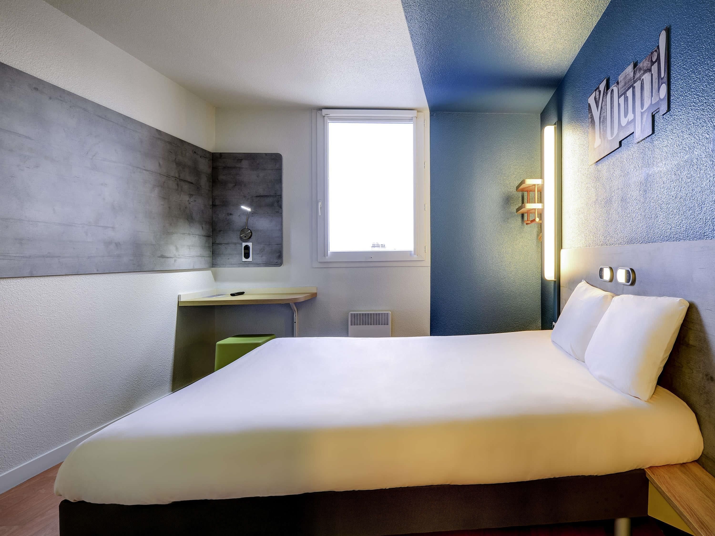 A modern hotel room with a bed, a desk, a window, and a wall-mounted light fixture.