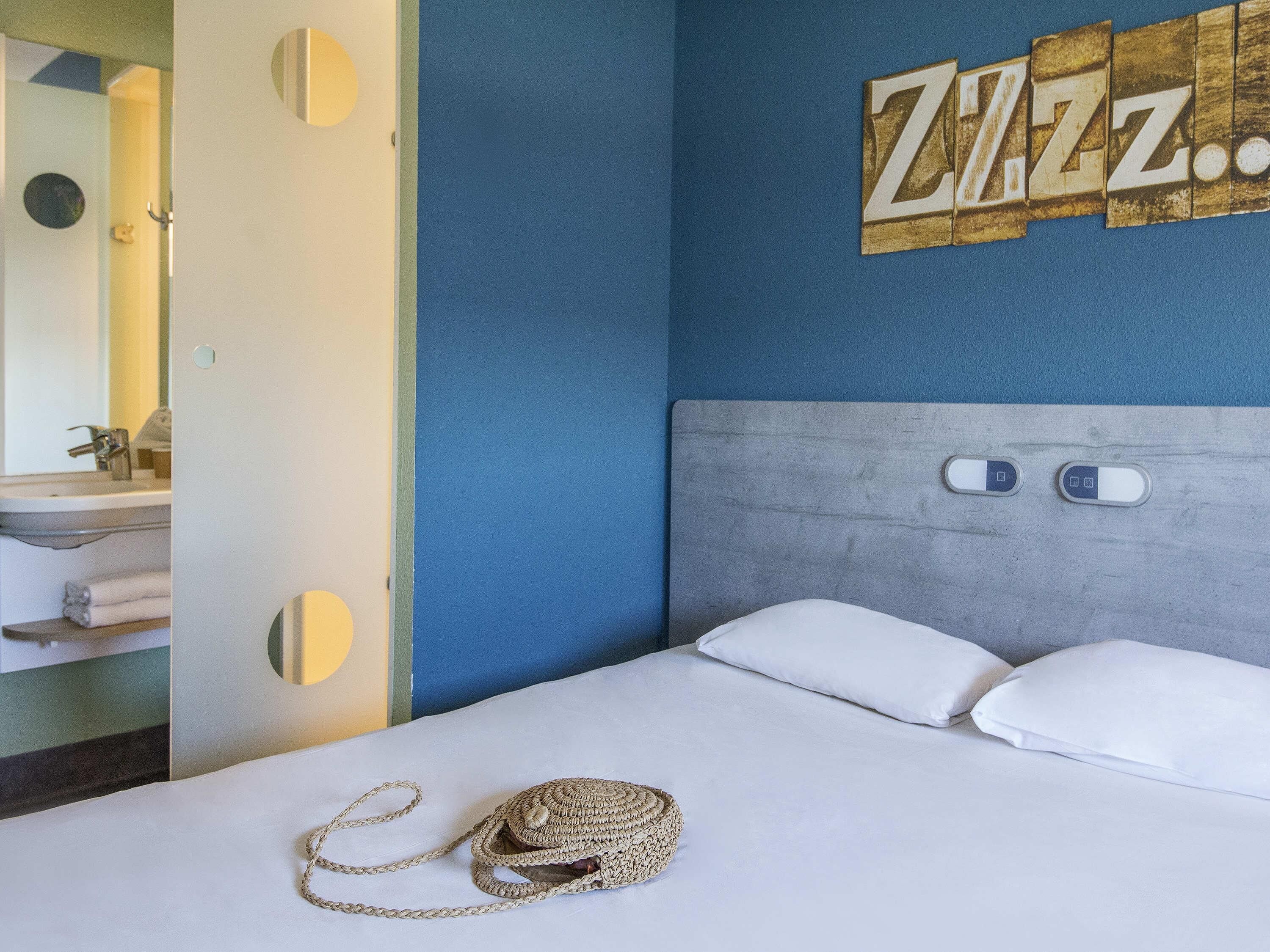 A hotel room with a bed, a headboard with two light switches, a wall with a 'Zzz' sign, and a bathroom visible through an open door.