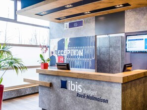 A reception desk with a sign for 'ibis budget Rueil-Malmaison,' a reception area with a large window, and a digital display on the wall. - ibis budget Rueil Malmaison (Nanterre)