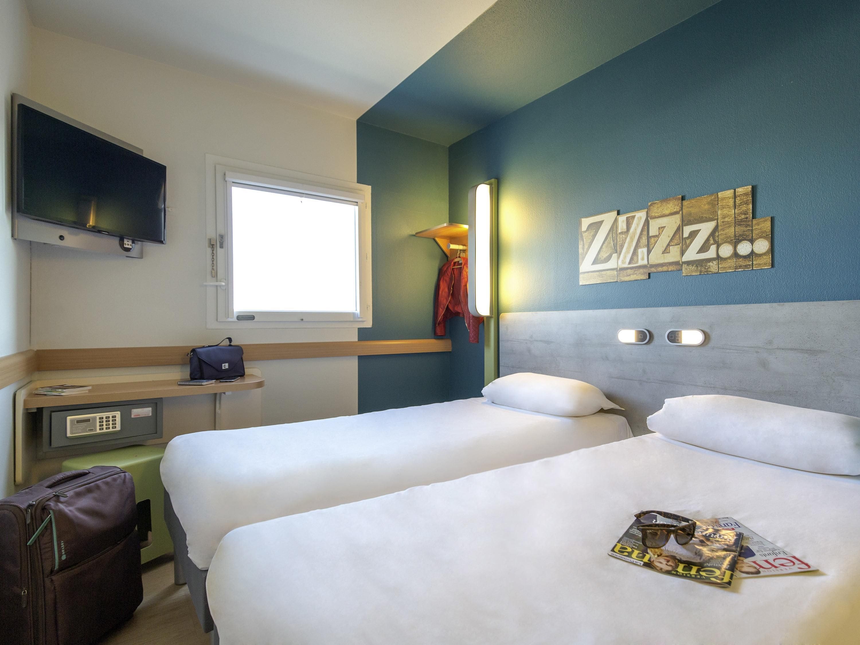 A hotel room with two beds, a desk with a chair, a TV mounted on the wall, and a window.