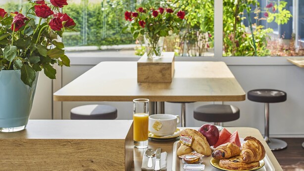 A breakfast table with croissants, a glass of orange juice, and a cup of tea, with a view of a garden through a window. - ibis budget Santeny (Santeny)