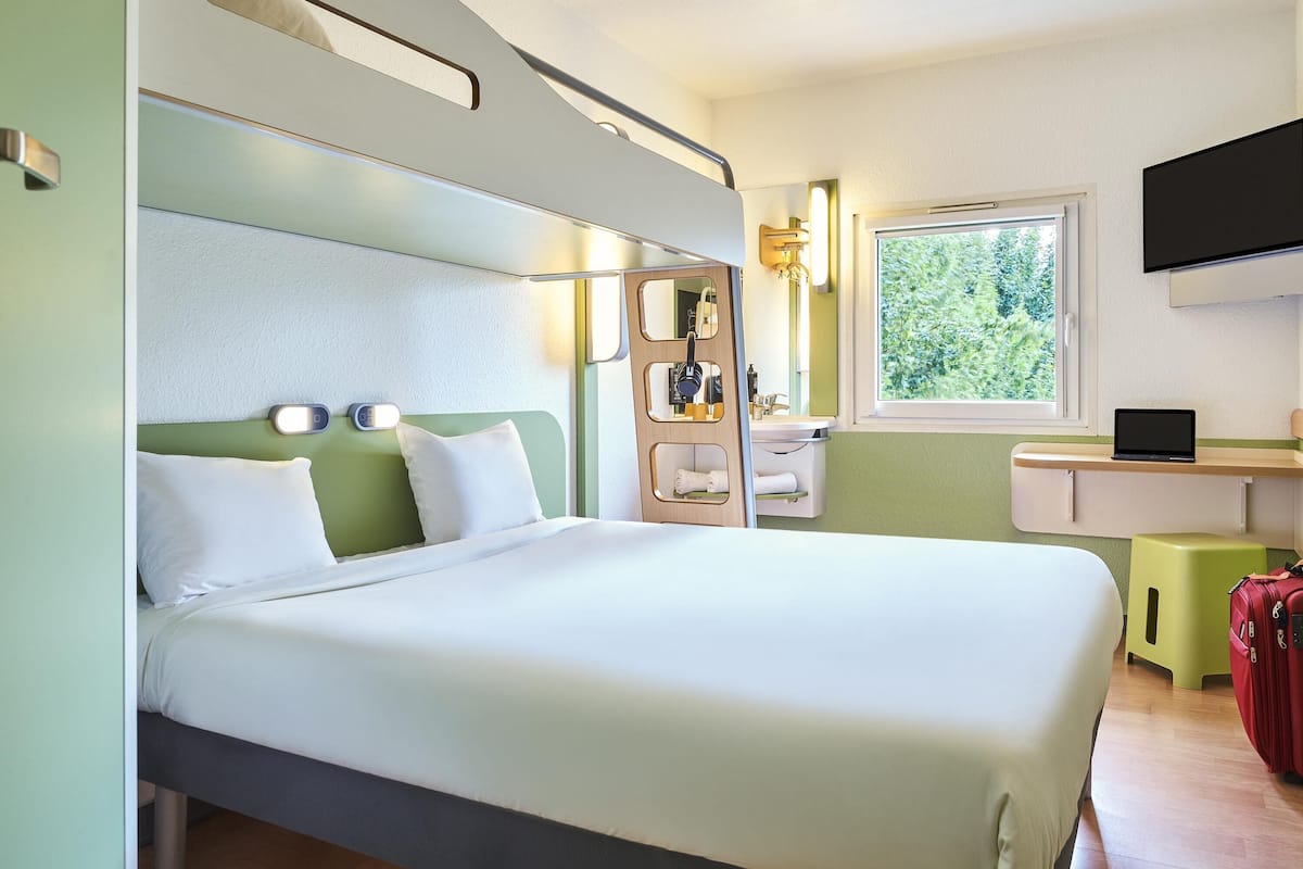 Habitación de hotel pequeña con cama, un escritorio, televisión y ventana con vistas a zonas verdes.