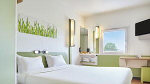 A hotel room with a bed, a bedside table, a mirror, a window, and a wall-mounted TV. - ibis budget Santeny (Santeny)