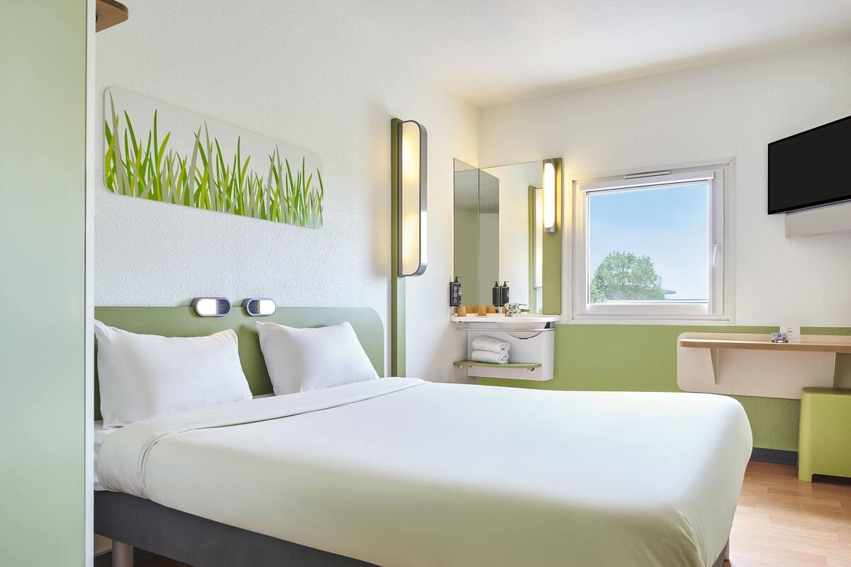 Habitación de hotel con una cama, una mesita de noche, un espejo, una ventana y un televisor montado en la pared.