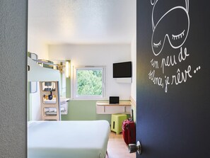 A modern hotel room with a bunk bed, a desk with a laptop, a window with a view of greenery, and a door with a decorative quote. - ibis budget Santeny (Santeny)