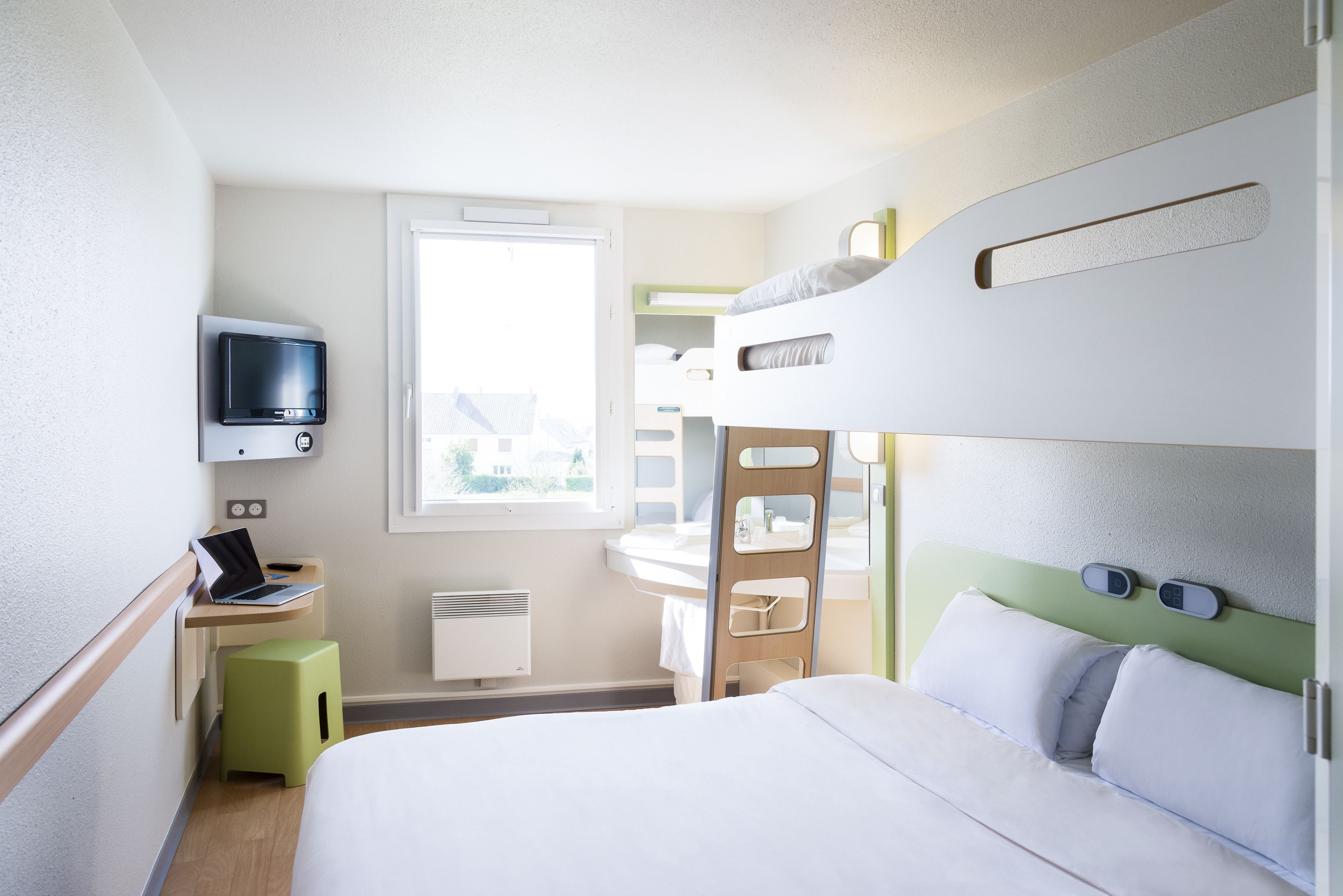 A compact hotel room with a bunk bed, a desk with a laptop, a TV, and a window providing natural light.