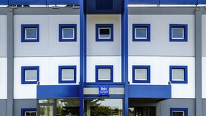Een modern hotel met een blauw-witte gevel, een opvallend logo en een zitgedeelte op straatniveau met groene stoelen.