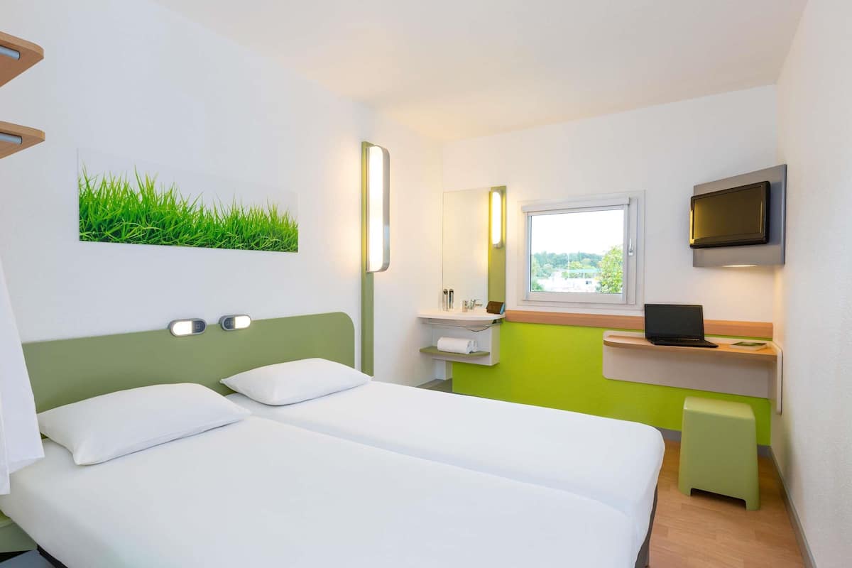 Une chambre d’hôtel avec un lit, un bureau avec un ordinateur portable, une télévision, une fenêtre et un mur vert.