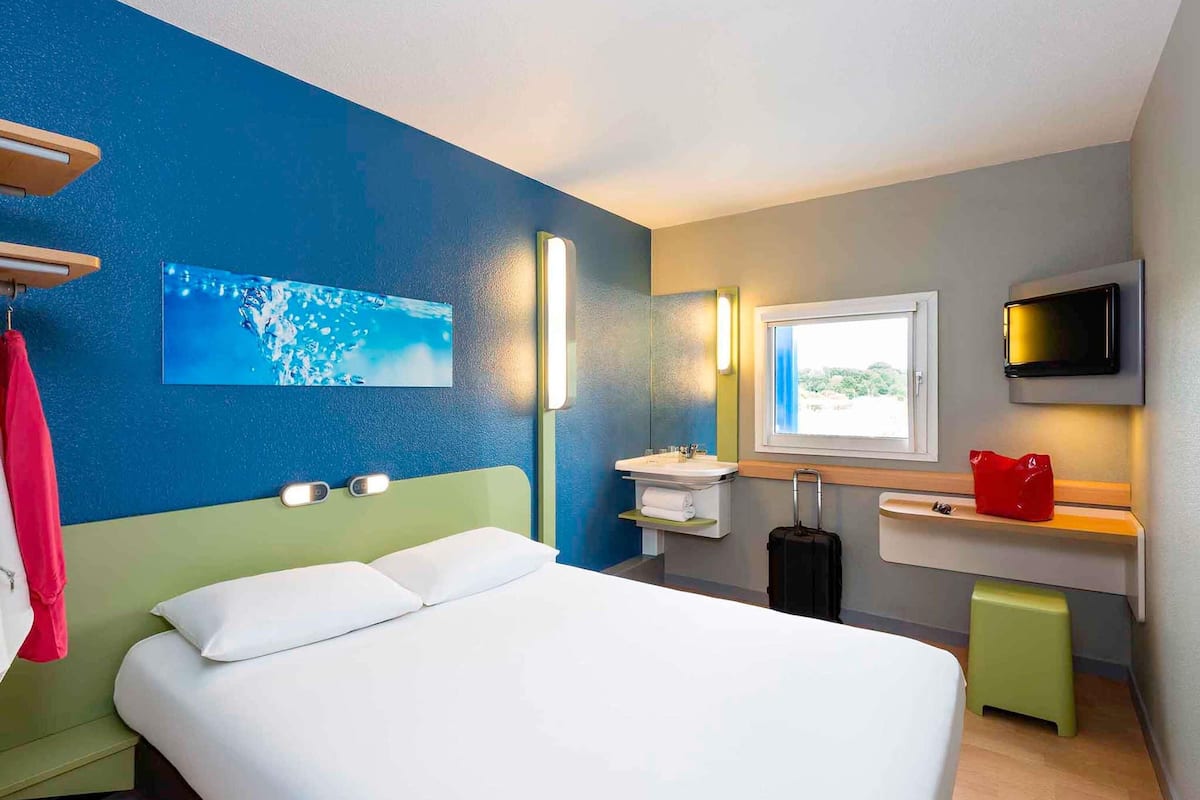 Une chambre d’hôtel avec un lit, un lavabo, un miroir, une fenêtre et un téléviseur.